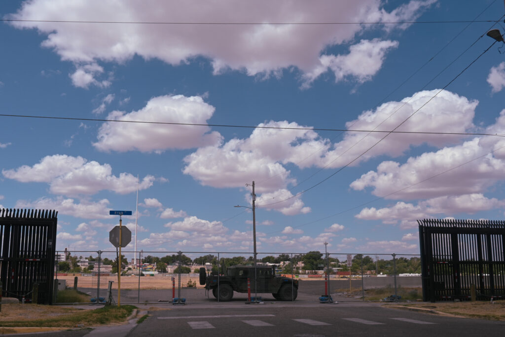 A military vehicle is parked longways across one of the entrance roads to access Shelby Park in Eagle Pass, Texas. In front of the vehicle is a chain link fence also blocking the entrance. On either side of the vehicle is the "border wall," a tall, black iron fence with thin slats.