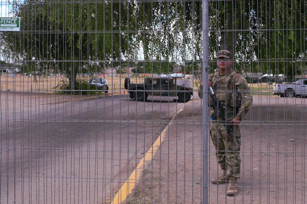 An military officer stands clutching an automatic rifle behind a fence. A military vehicle and two other law enforcement vehicles are behind the officer. This is another entry point into Shelby Park, which was closed to visitors.